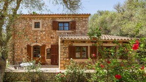 A stone house with a tiled roof, a patio with a table and chairs, and a garden with red flowers.