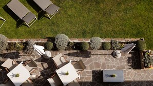 Une terrasse extérieure avec un sol en pierre, des tables et des chaises blanches, et des haies soigneusement taillées.