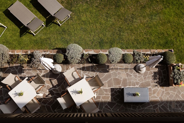 An outdoor patio with stone flooring, white tables and chairs, and neatly trimmed hedges.