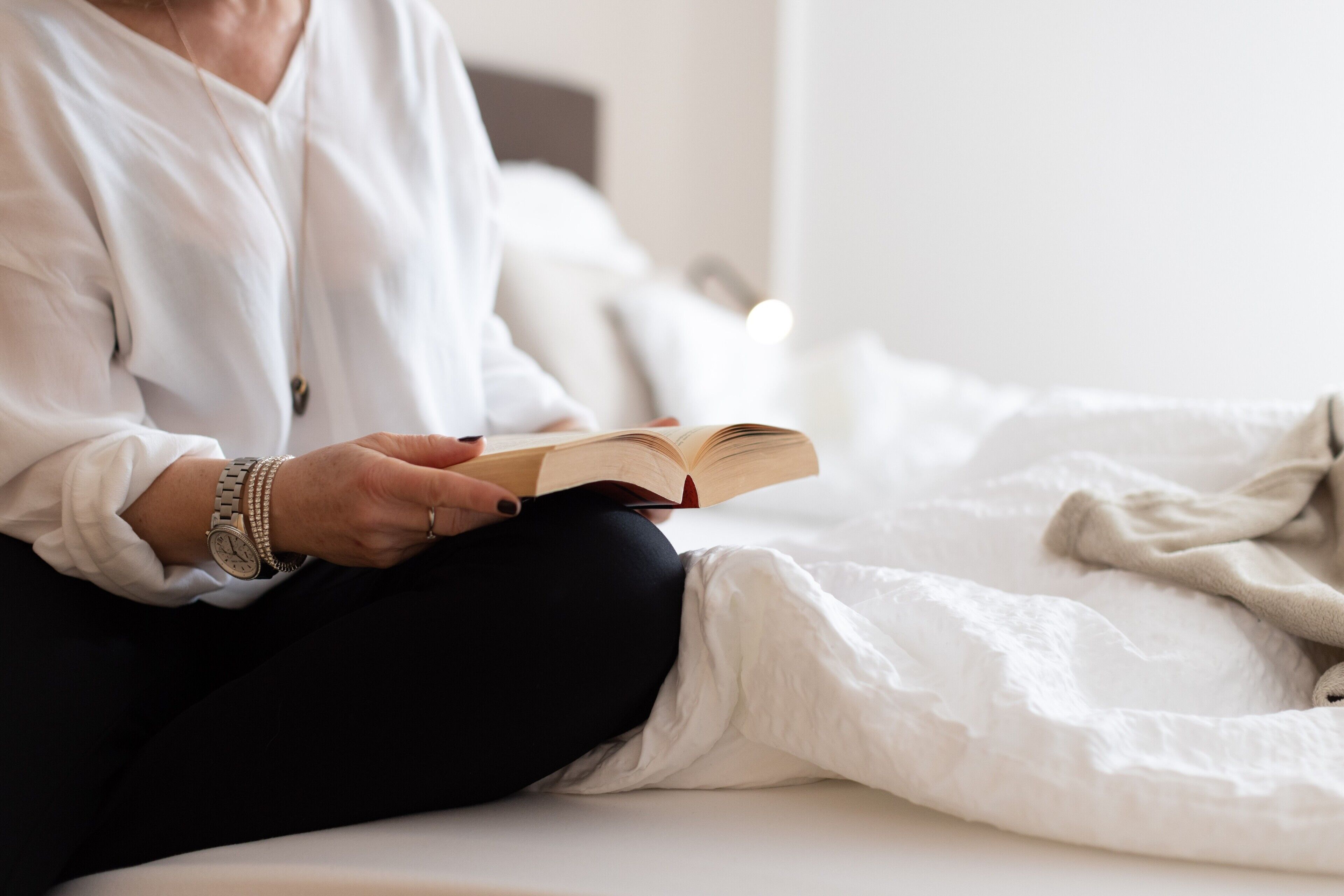 A person sitting on a bed with a book, wearing a white shirt and black pants.