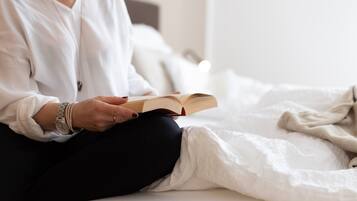 Una persona sentada en una cama leyendo un libro, vestida con una camisa blanca y pantalones negros.