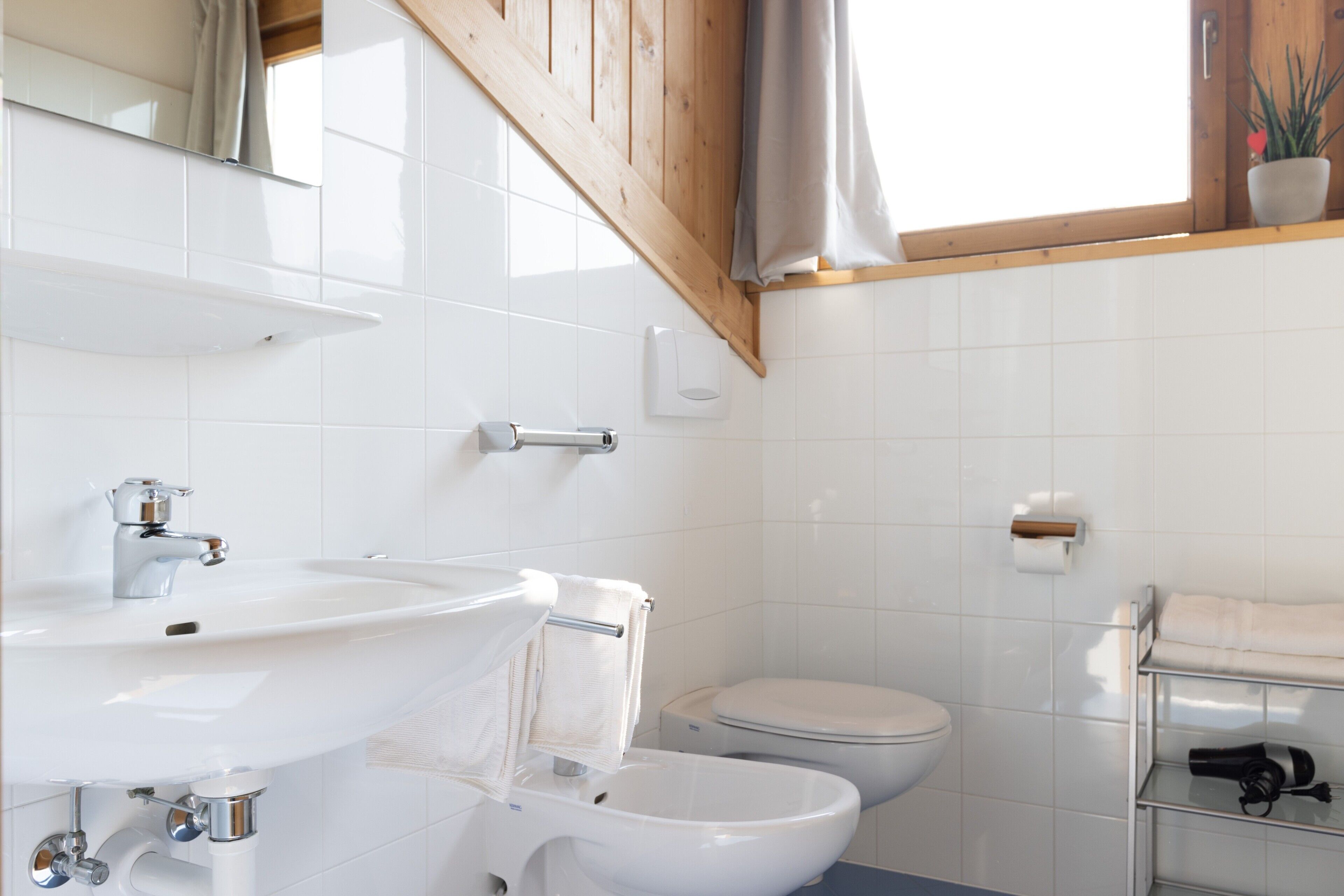 A bathroom with a white sink, toilet, and bidet, wooden ceiling beams, and a potted plant on the window sill.
