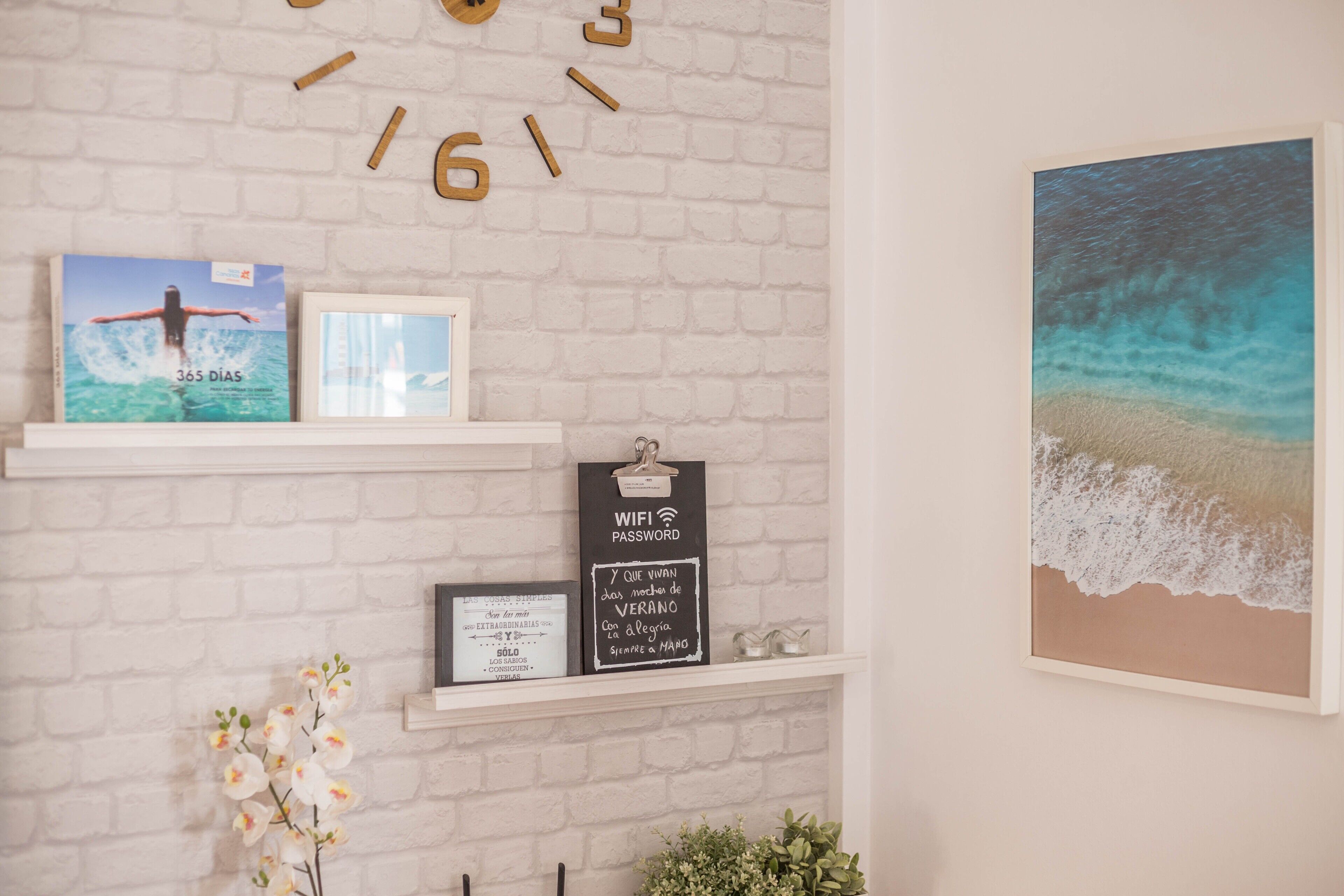 A white brick wall with shelves displaying framed pictures, a clock, and a beach scene painting.
