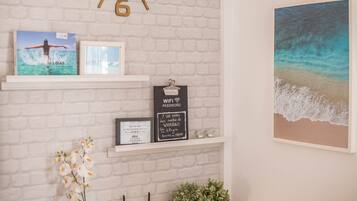 A white brick wall with shelves displaying framed pictures, a clock, and a beach scene painting.