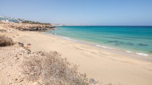A beach with clear turquoise water, sandy shore, and a building with multiple balconies in the background. - Maritime Holiday Home Casa Yerida with Pool and Wi-Fi (Cotillo)