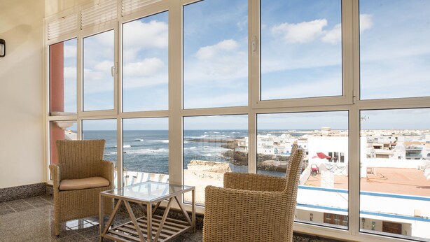 A room with large windows, wicker furniture, and a view of the sea and buildings. - Maritime Holiday Home Casa Yerida with Pool and Wi-Fi (Cotillo)