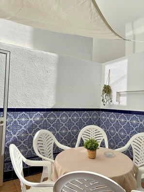 A corner room with white walls, blue patterned tiles, white plastic chairs, a round table with a beige tablecloth, and a hanging plant.