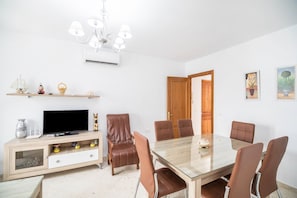 A dining area with a glass table, six chairs, a television on a wooden cabinet, and a chandelier.