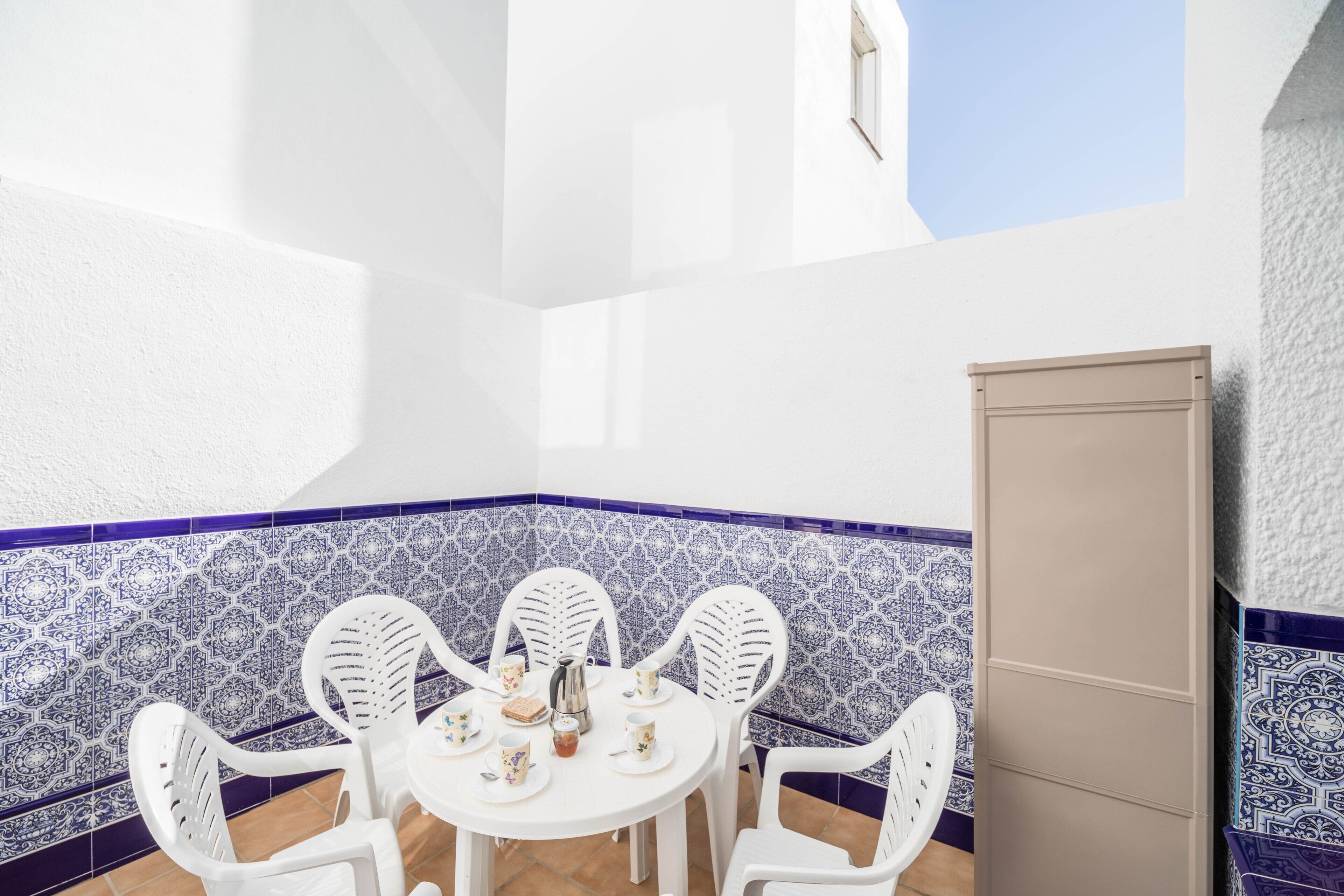 Un patio en esquina con una mesa y sillas blancas, un revoque de azulejos y un armario color arena.