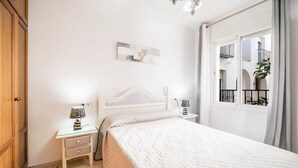 A bedroom with a bed, bedside tables, a nightstand, a wall-mounted artwork, and a window with a view of balconies. - Newly Renovated Apartment “Conil Centro” with Wi-Fi, Terrace & Air Conditioning; Parking Available (Conil de la Frontera)