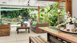 A covered outdoor area with wicker furniture, a wooden table, and a view of lush greenery. - Luxurious Family Villa with Private Pool, Nestled in Cairns' Tropical Paradise (Whitfield)
