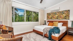 A bedroom with a large window, a bed with multiple pillows, a ceiling fan, and a wall-mounted air conditioner. - Luxurious Family Villa with Private Pool, Nestled in Cairns' Tropical Paradise (Whitfield)