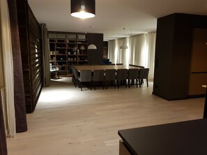 Een ruime woonkamer met een houten vloer, een eettafel met stoelen, een boekenkast en een keukenhoek.