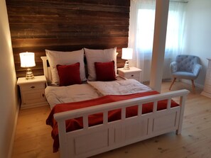 Een slaapkamer met een houten hoofdbord, wit beddengoed, rode kussens, twee nachtkastjes met lampen en een grijze fauteuil.