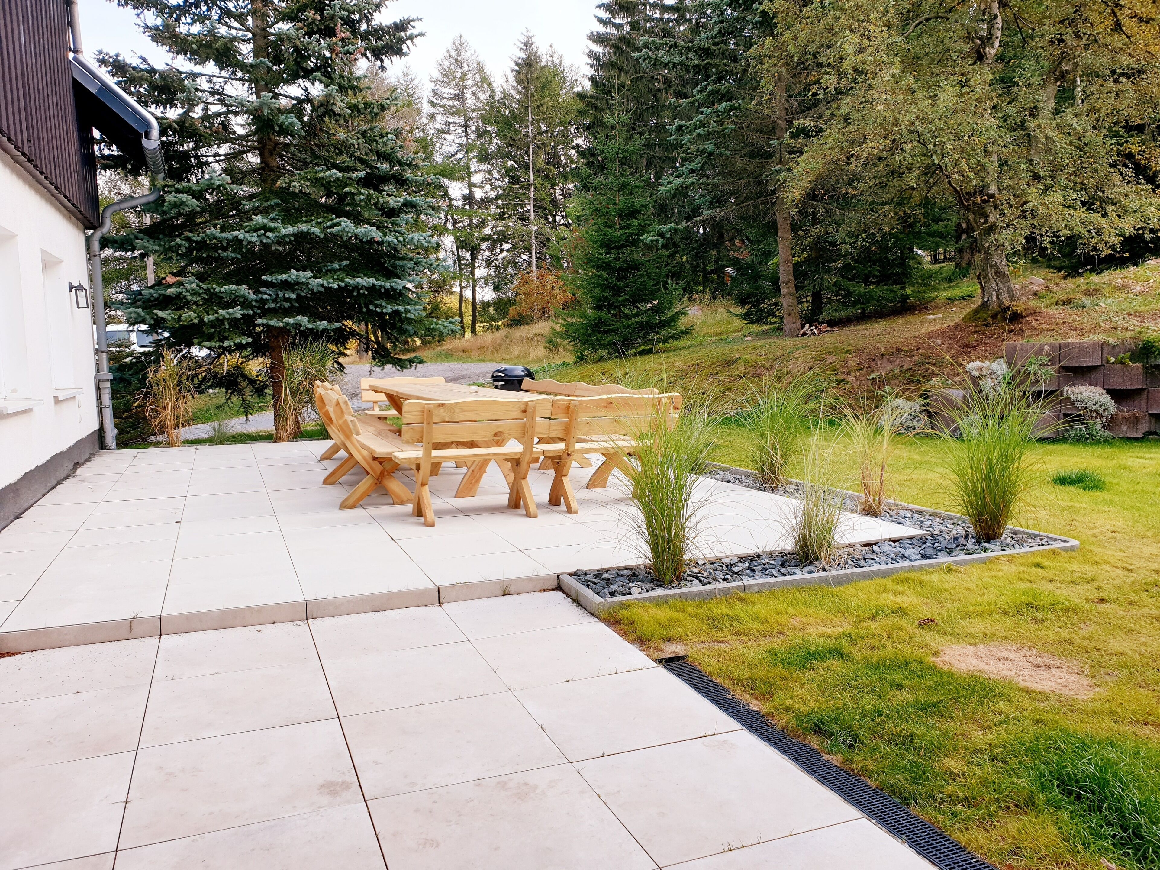 A patio with a wooden table and chairs, surrounded by a garden with grass and small plants.