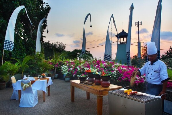 An outdoor dining area with tables set for a meal, a chef cooking on a grill, and decorative arches in the background.