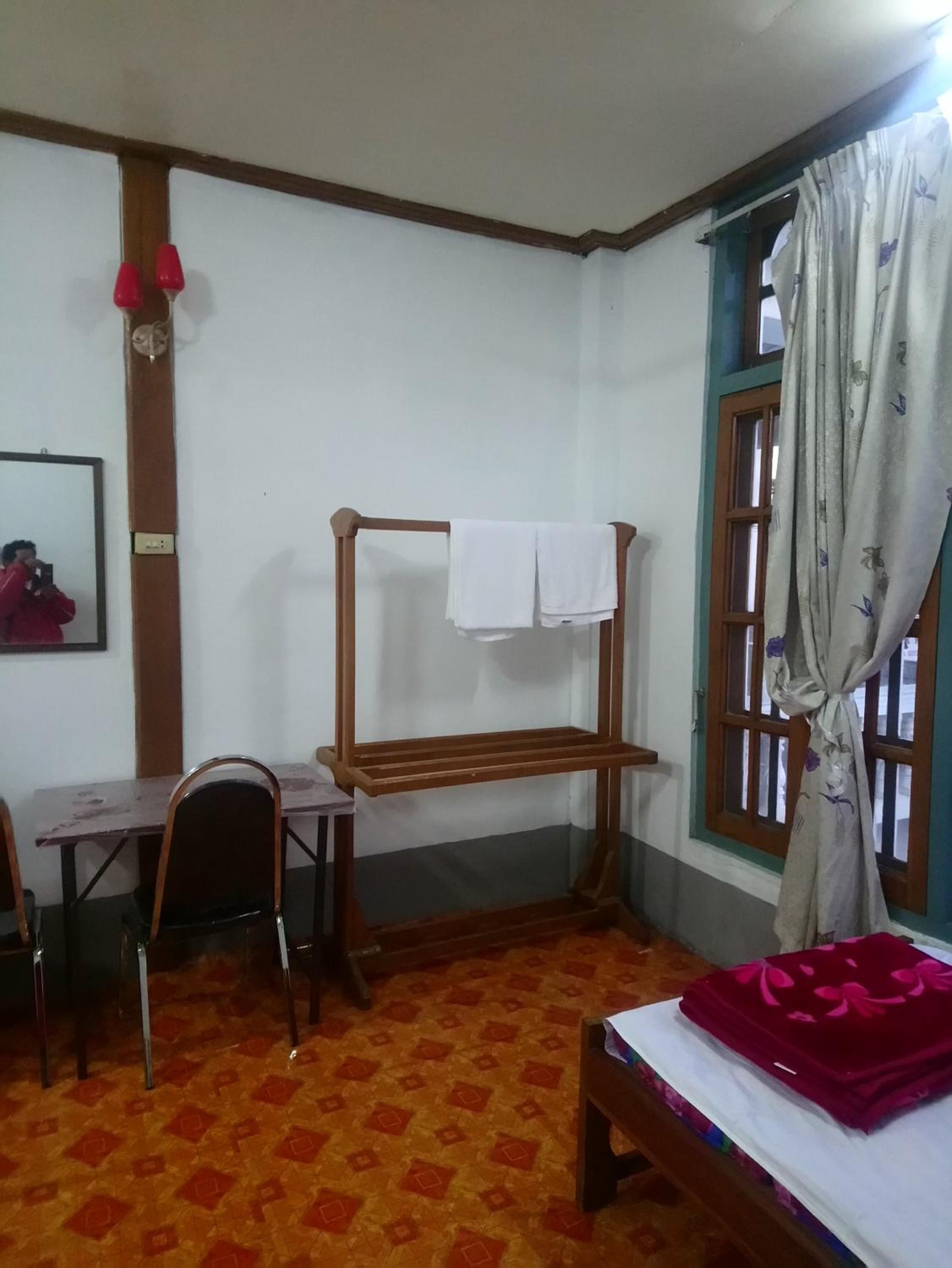 Ein Zimmer mit einem Bett, einem Schreibtisch mit Stuhl, einem Holzregal mit Handtuch, einem Fenster mit Vorhängen und einem gemusterten Boden.