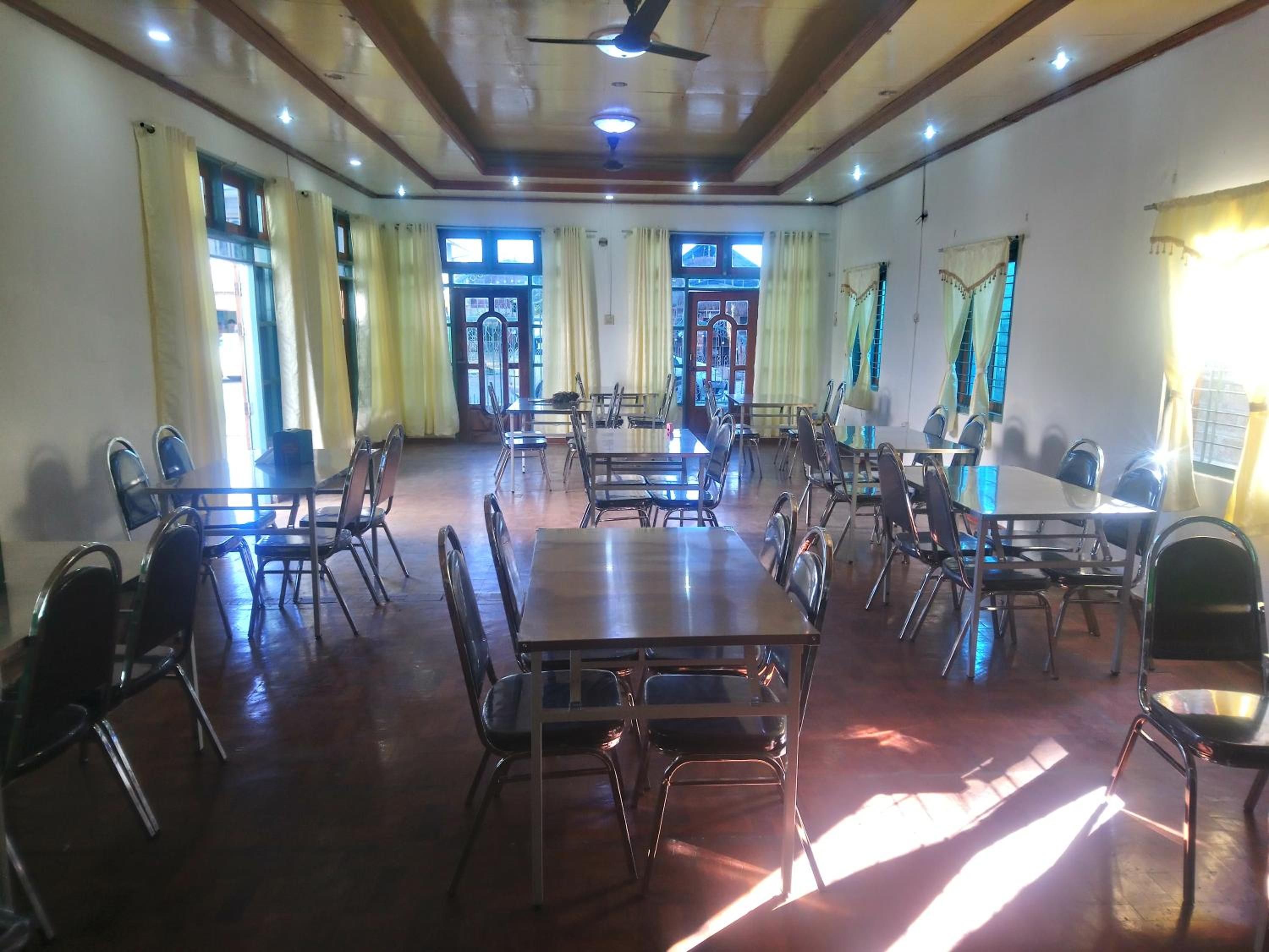 Une salle à manger spacieuse, dotée de nombreuses tables et chaises, de grandes fenêtres avec des rideaux et d’un ventilateur de plafond.