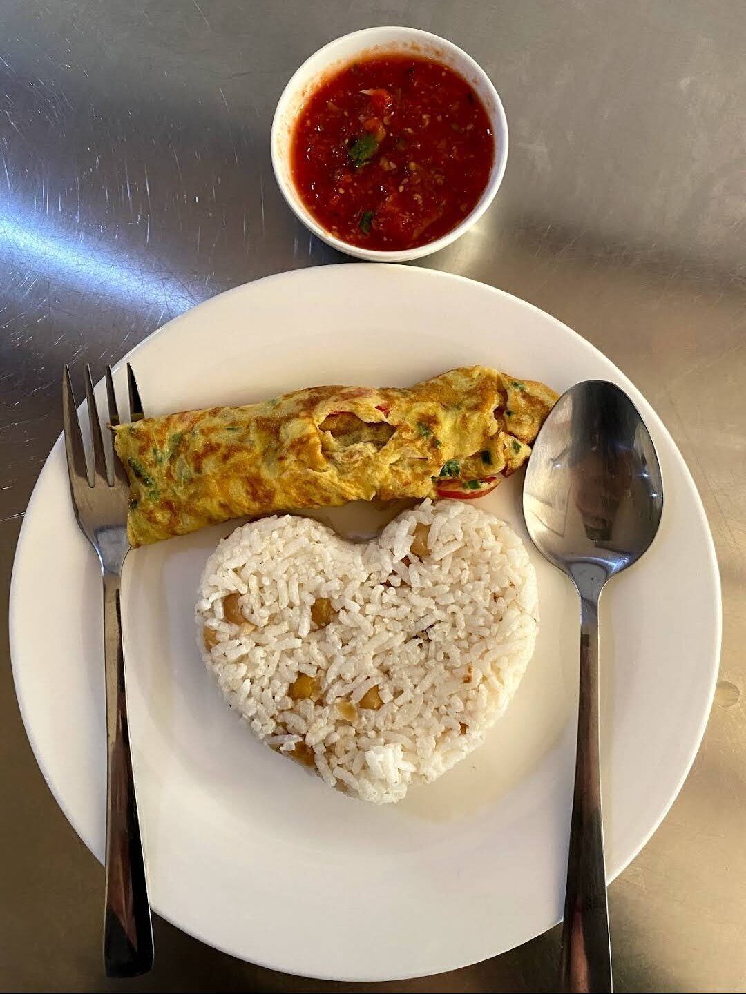 En tallerken med hvid ris og omelet, ledsaget af en lille skål med rød sauce og bestik på et metalbord.