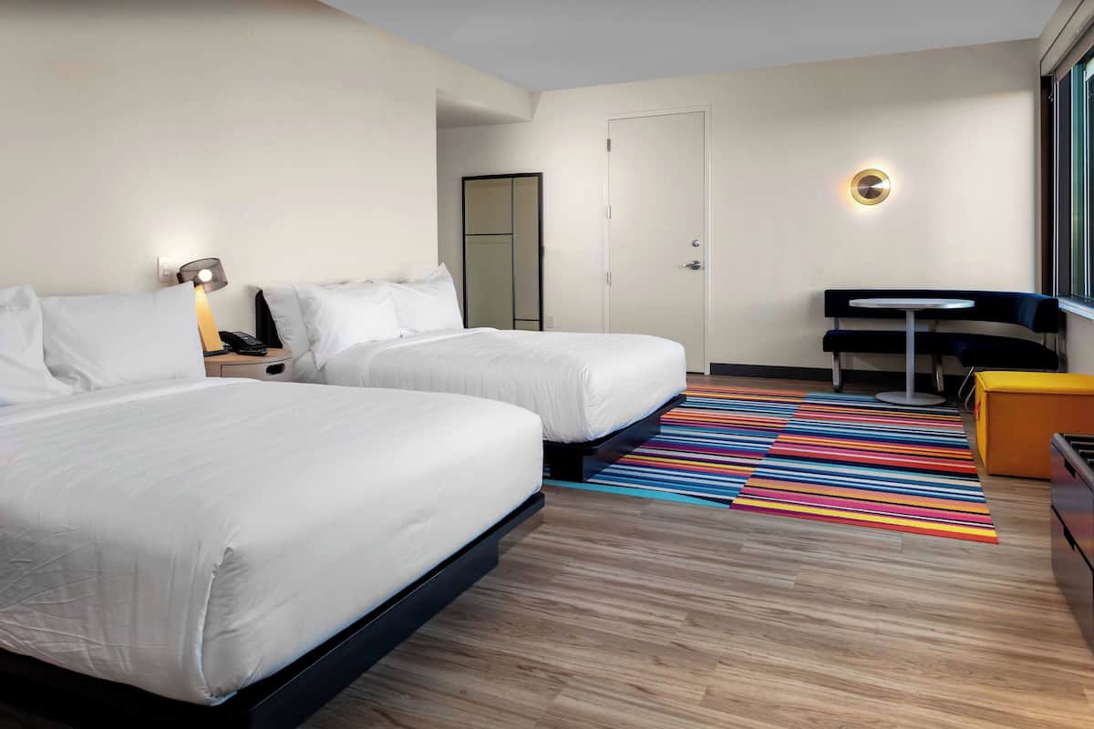 Ein Hotelzimmer mit zwei Betten, einem kleinen Tisch, einem Stuhl, einem Spiegel und einem farbenfrohen Streifen Teppich.