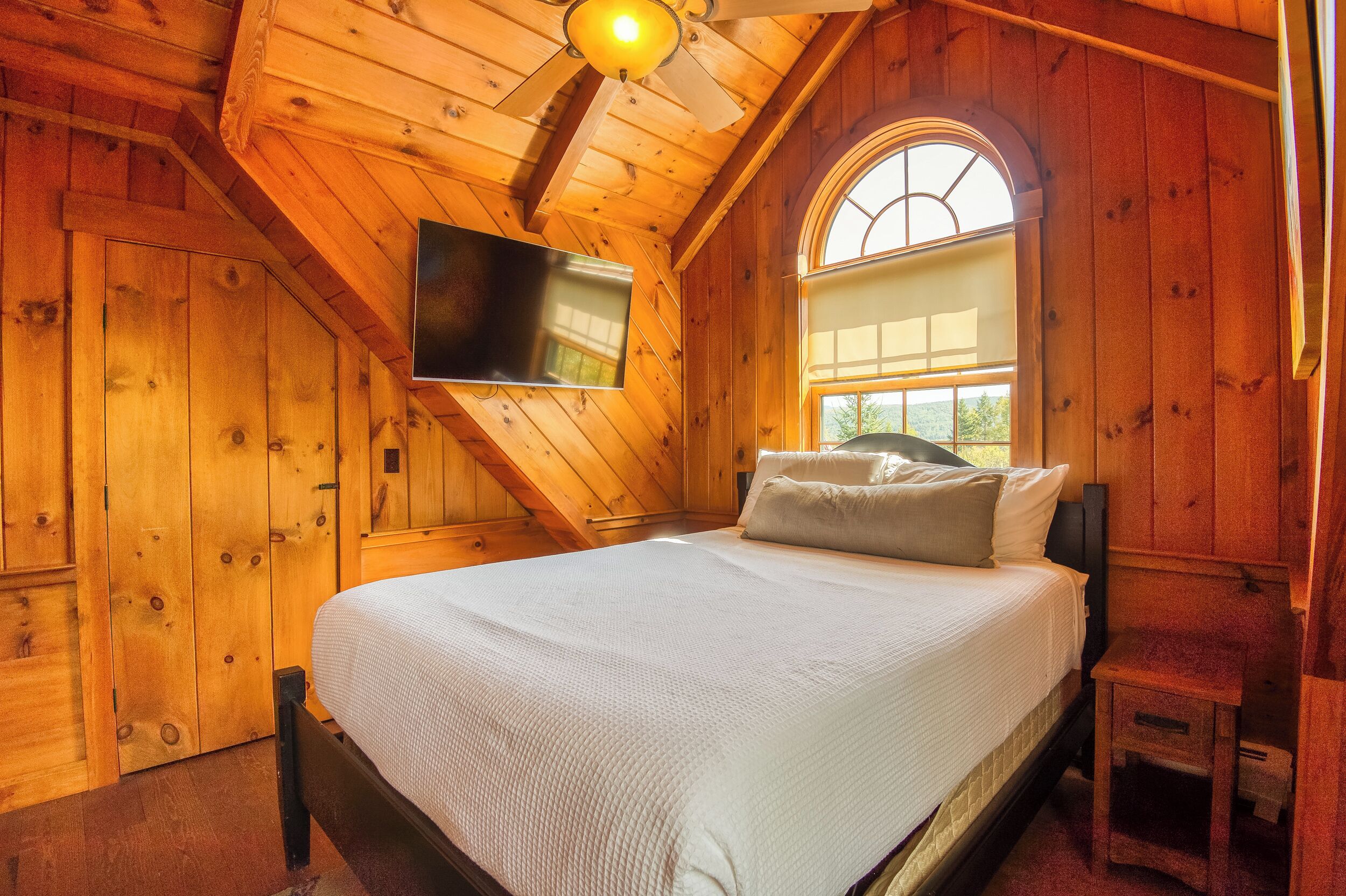Un dormitorio de cabaña de madera con una cama, un televisor montado en la pared y una ventana con vistas a follaje.