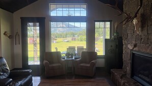 A cozy living room with a fireplace, large windows, and a view of a green landscape. - Country setting close to Glacier National Park (Columbia Falls)