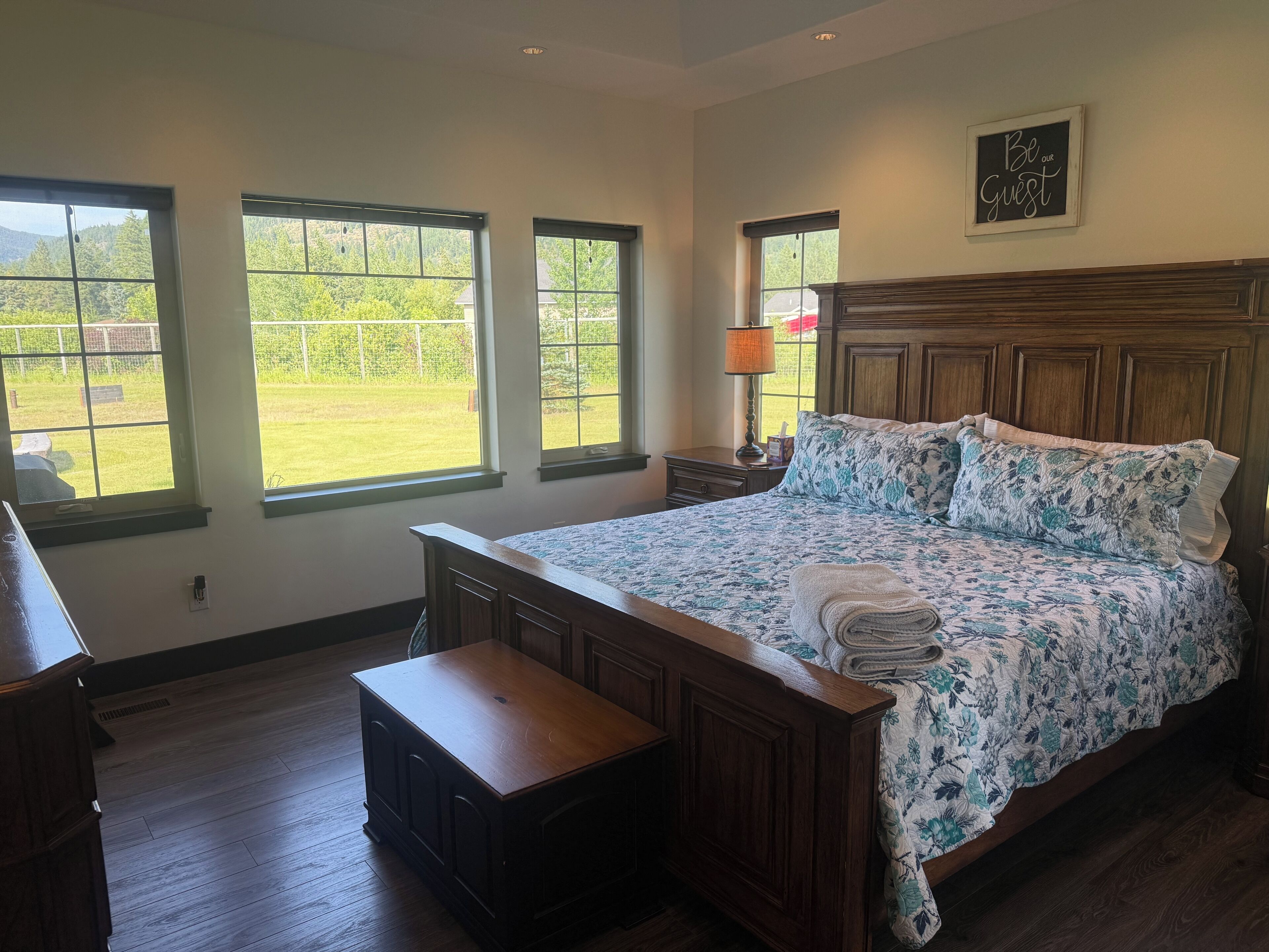 Un dormitorio con una cama grande, cabecera de madera y vista a un jardín con césped a través de tres ventanas.