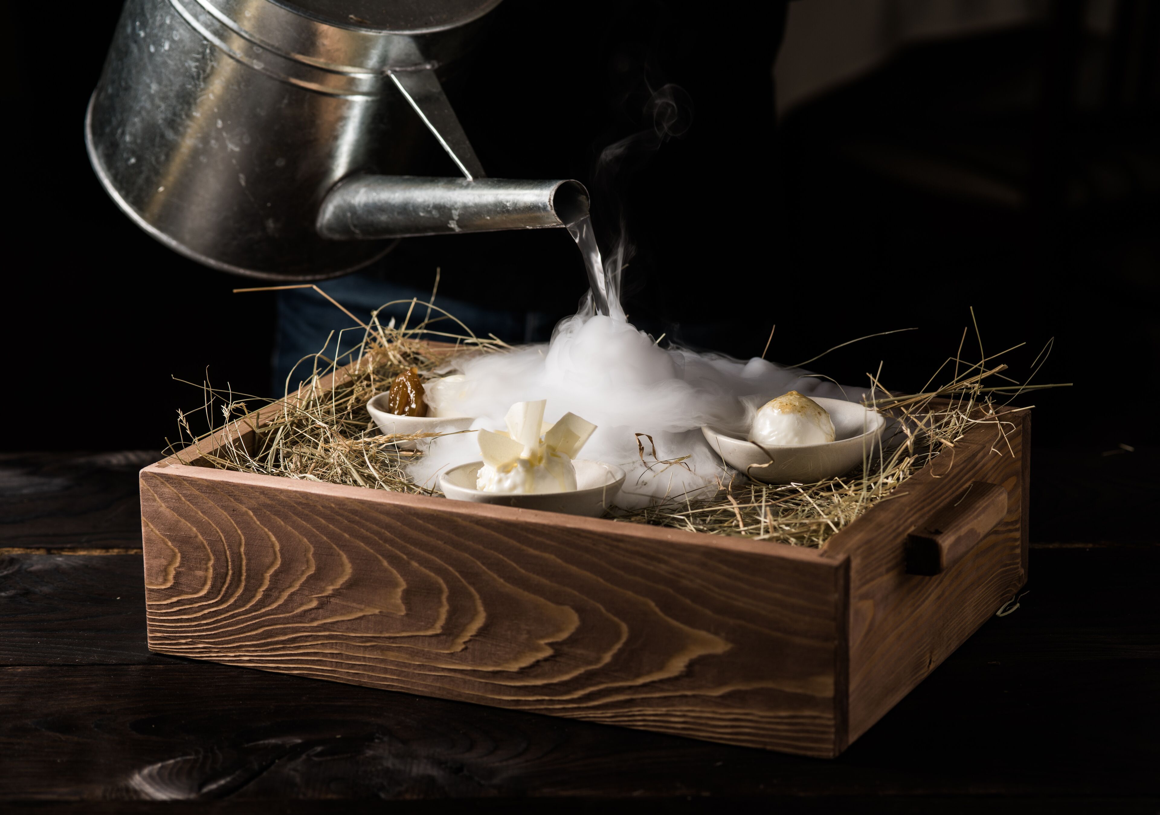 A wooden box with a metal pot pouring water onto a bed of straw, surrounded by bowls and flowers.