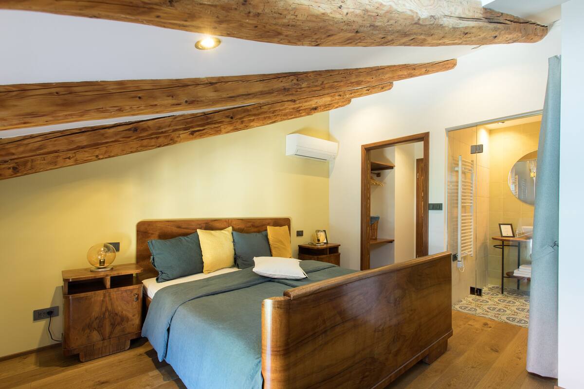 A bedroom with a wooden bed, a nightstand, and a bedside table. There is a wooden ceiling beam and an air conditioning unit on the wall.