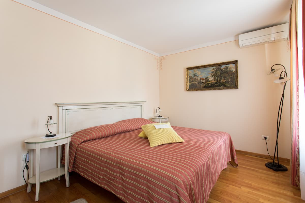 A bedroom with a bed, bedside table, lamp, and an air conditioning unit.