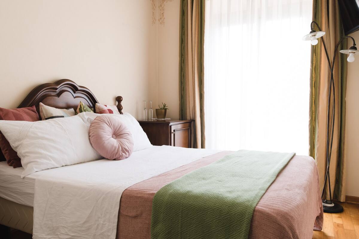 A neatly made bed with a wooden headboard, a bedside table with a lamp, and a window with curtains.