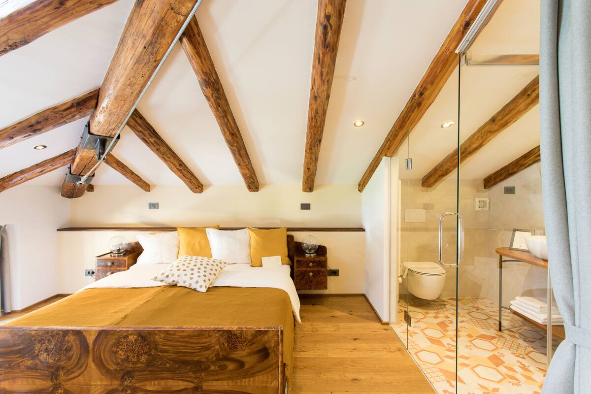 A bedroom with a wooden ceiling, a bed with a wooden headboard, a nightstand, and a glass shower enclosure.