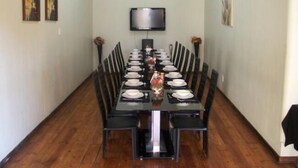 A dining area with a long table set for a meal, flanked by chairs, a television mounted on the wall, and framed artwork on the walls. - Times Premier Lodge (Vryburg)