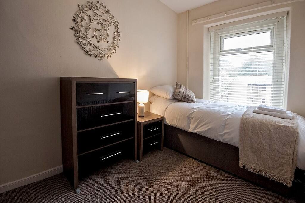 Ein Schlafzimmer mit Bett, Schminktisch, Nachttisch, Wandbild und einem Fenster mit Jalousien.