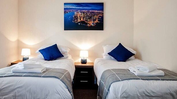 Una camera d'albergo con due letti, ciascuno con un cuscino e una lampada. Sopra i letti, un quadro incorniciato appeso al muro.