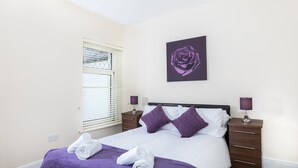 Una cama bien hecha con una manta y almohadas moradas, una mesita de noche con una lámpara y una rosa morada enmarcada en la pared.