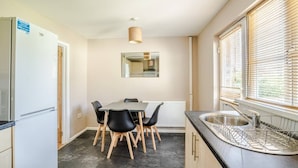 A compact kitchen with a dining table, chairs, a refrigerator, a sink, and a window with blinds. - Twyn Lodge - 3 Bed House (Pontlliw)