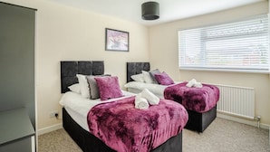 A bedroom with two beds, each with a dark headboard, a window with blinds, and a framed picture on the wall. - Twyn Lodge - 3 Bed House (Pontlliw)