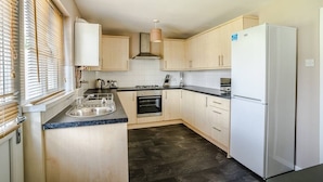A compact kitchen with wooden cabinets, a built-in oven, a stainless steel refrigerator, and a window with blinds. - Twyn Lodge - 3 Bed House (Pontlliw)