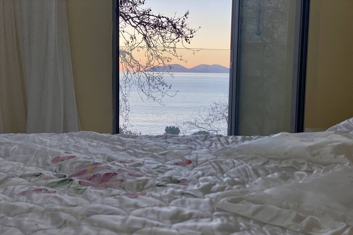 Een slaapkamer met een bed, een raam met uitzicht op zee en bergen, en een gordijn.