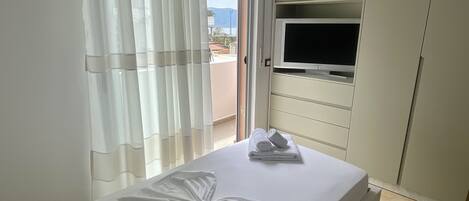 A neatly made bed with a white towel and a rolled towel on it, a television on a shelf above the bed, and a view of the outside through a window with curtains.