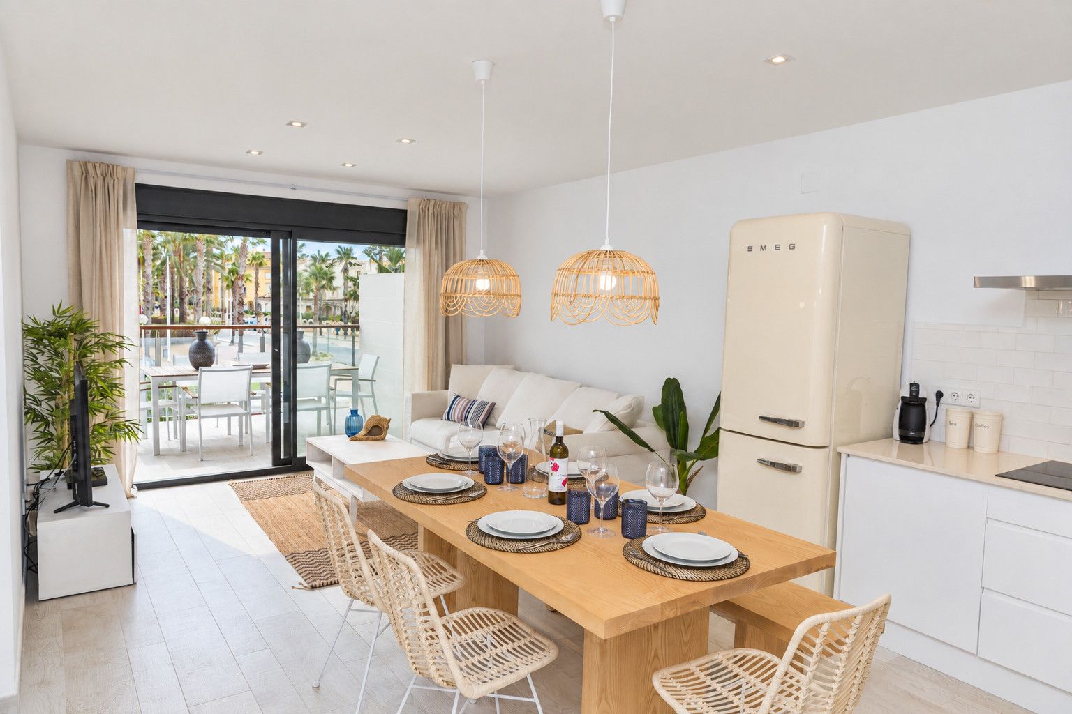 Una cocina moderna con armarios blancos, una mesa de comedor de madera preparada para cuatro personas y un amplio ventanal con vistas a un área exterior tropical.