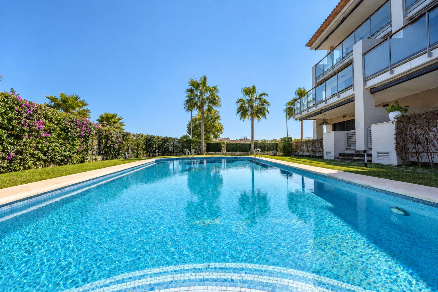 A clear swimming pool with a tiled edge, surrounded by lush greenery and palm trees.