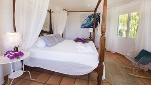 Een hemelbed met witte lakens, een kleine ronde tafel met een lamp en bloemen, en een schommelstoel in een lichte kamer met uitzicht op bomen door het raam.