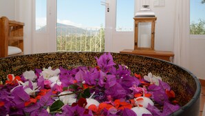 Een kamer met een grote decoratieve schaal vol bloemen, een houten bankje, een witte deur en uitzicht op een balkon met een leuning en een berg op de achtergrond.