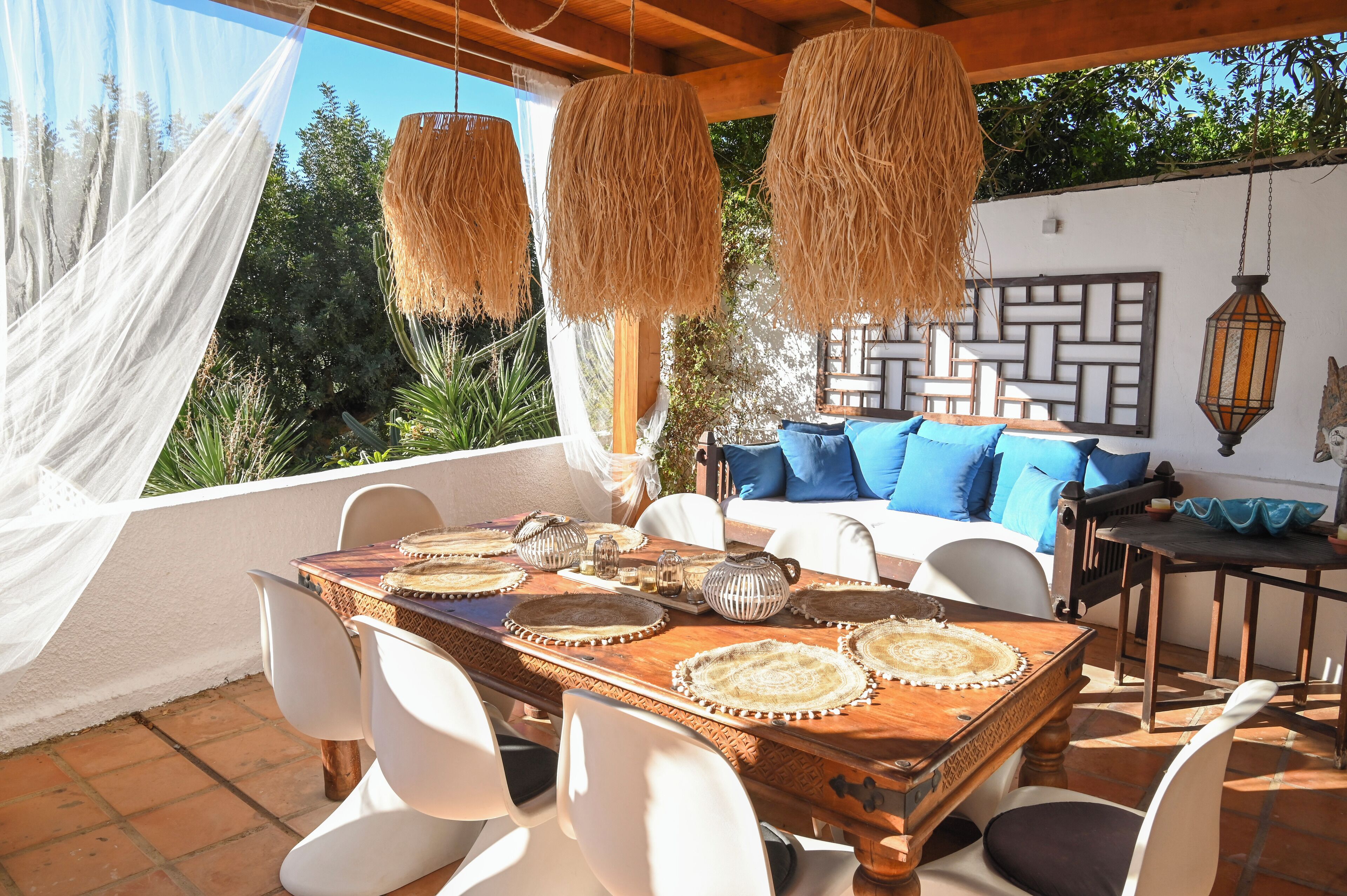 A patio with a wooden table set for dining, white chairs, and hanging rattan lamps.