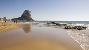 A beach with a prominent rock formation in the background, a sandy shore, and a few people walking. - Romantic loft for couples with views of the ocean, the salt flats, and the natural park (Calpe)