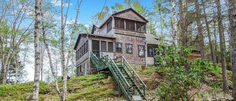 A rustic cabin with a brick exterior, a green staircase leading to the entrance, surrounded by a forested area.