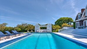 A swimming pool with lounge chairs and a covered seating area, surrounded by greenery and a building with a staircase. - Elegant Watefront home with Oversized Gunite Heated Pool, Tennis, Living... (Water Mill)