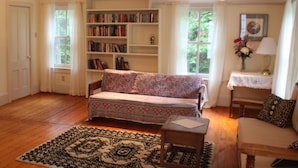 A cozy living room with a patterned sofa, a bookshelf filled with books, and a wooden floor. - Classic House on Peters Cove (Blue Hill)